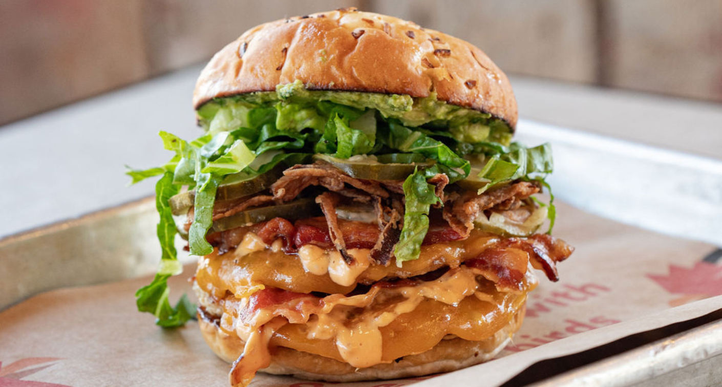 Sandwich with guacamole, lettuce, pickles, bacon, and chicken patties on a tray.
